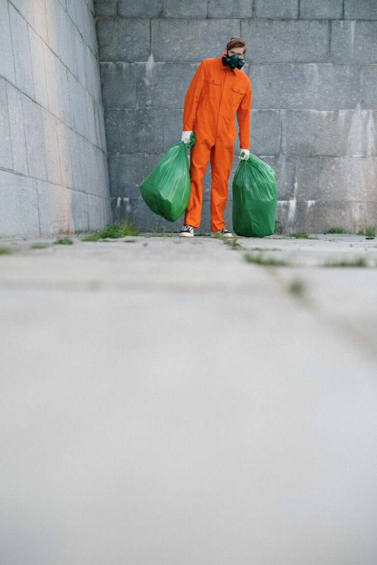 A waste collector in an orange suit holds green trash bags against a concrete wall backdrop.